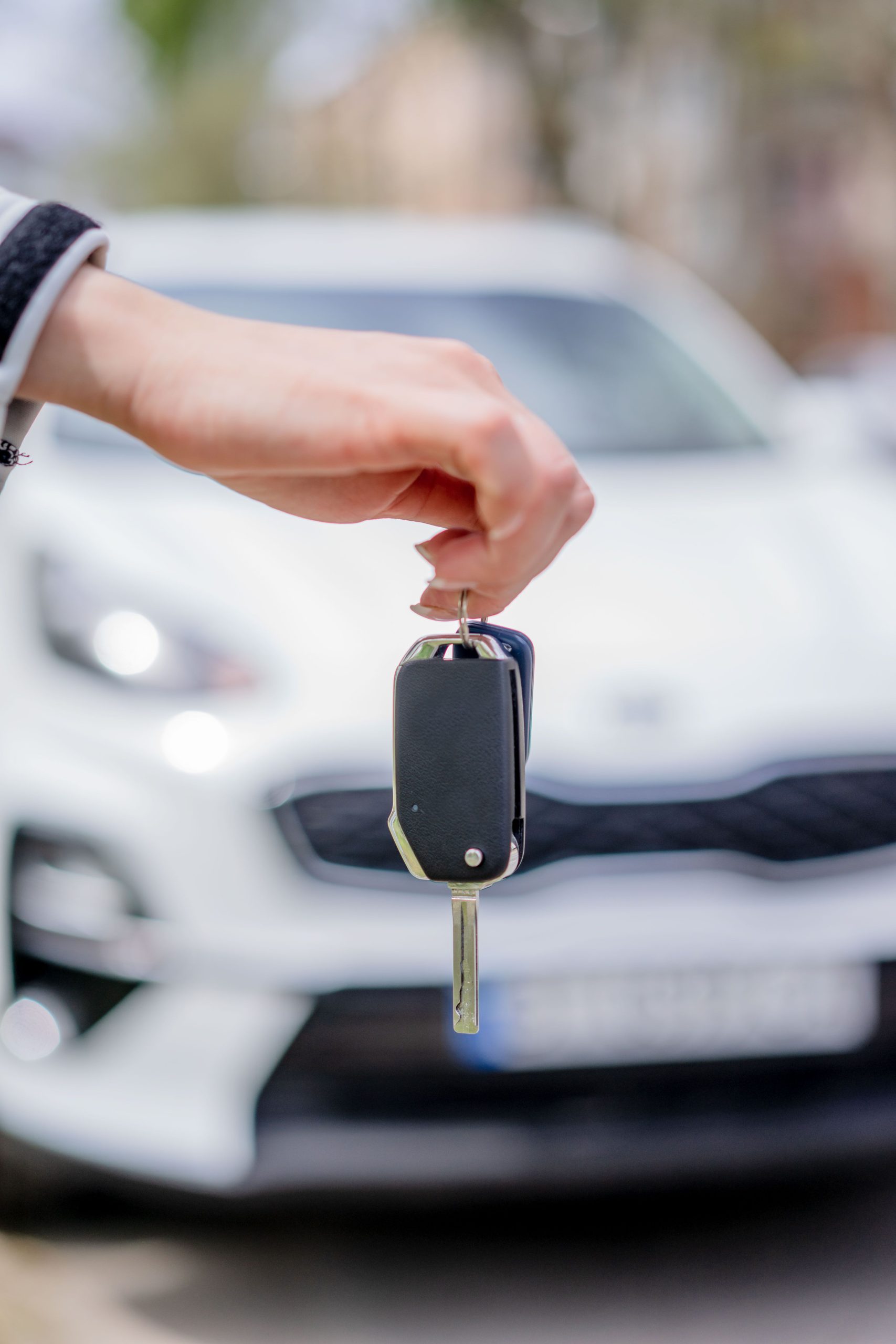 Hand holding a car key in front of a parked white car